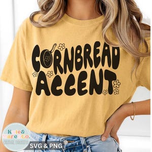 Könnte beinhalten: Senfgelbes T-Shirt mit dem Schriftzug "CORNBREAD ACCENT" in großer, schwarzer Retro-Schrift. Das Design beinhaltet eine Pfannengrafik und Blumenakzente. Ein lässiges Kleidungsstück für den Alltag.