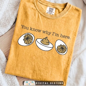 Puede incluir: Una camiseta de manga corta color amarillo mostaza con el texto negro 'You know why I'm here.' sobre un gráfico de tres huevos duros cortados por la mitad. El huevo central tiene un relleno marrón, mientras que los dos exteriores muestran yemas texturizadas.