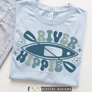May include: A light blue t-shirt featuring a graphic design with a dark blue kayak and paddle. The text "RIVER HIPPIE" is printed in a wavy, retro-style font in shades of blue and sage green, accented by small flower and star icons.