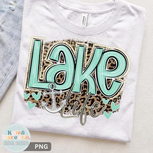 May include: A white t-shirt featuring a graphic design with the text "Lake life" in mint green with white stripes. The text is layered over a brown and black leopard print background, accompanied by a silver anchor and small mint green hearts.
