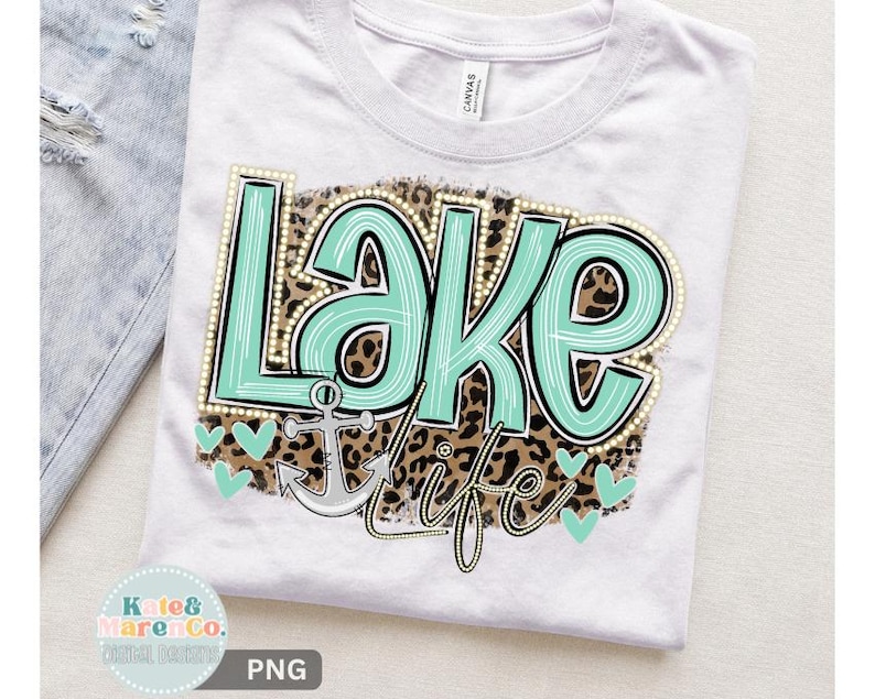 May include: A white t-shirt featuring a graphic design with the text "Lake life" in mint green with white stripes. The text is layered over a brown and black leopard print background, accompanied by a silver anchor and small mint green hearts.