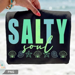 Pu&ograve; includere: Telo mare nero con le parole "SALTY soul" in verde acqua e verde. L'asciugamano presenta illustrazioni di conchiglie e stelle marine lungo il bordo inferiore. L'asciugamano &egrave; tenuto da una persona con un braccialetto turchese e dorato.