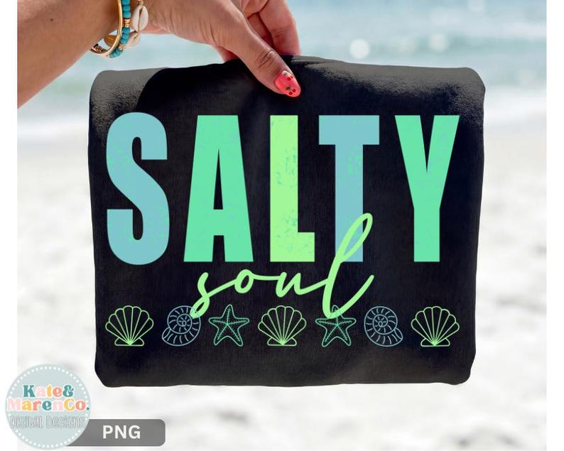 Pu&ograve; includere: Telo mare nero con le parole "SALTY soul" in verde acqua e verde. L'asciugamano presenta illustrazioni di conchiglie e stelle marine lungo il bordo inferiore. L'asciugamano &egrave; tenuto da una persona con un braccialetto turchese e dorato.