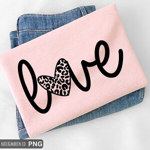 May include: A light pink t-shirt with the word "love" in black script. The "o" in love is replaced with a heart shape filled with a leopard print pattern. The shirt is folded and placed on top of a pair of blue jeans. The text "SVG KATE & MAREN CO PNG" is at the bottom.