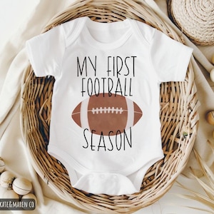 May include: White baby bodysuit featuring the text "MY FIRST FOOTBALL SEASON" and a brown American football graphic. The bodysuit is presented in a woven basket with decorative items.