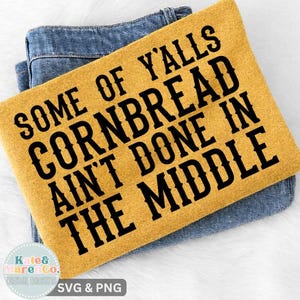 May include: Mustard yellow rectangular item with the text "SOME OF Y'ALLS CORNBREAD AIN'T DONE IN THE MIDDLE" in black. The item is resting on top of a pair of blue jeans. The bottom left corner has a logo that says "Kate & Marenco."