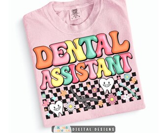 Op de afbeelding: Een lichtroze t-shirt met de tekst DENTAL ASSISTANT in kleurrijke letters in bubbelstijl. Onder de tekst staat een zwart-wit dambordpatroon versierd met cartoon-tanden, een tandartsstoel en kleine kleurrijke bloemen.