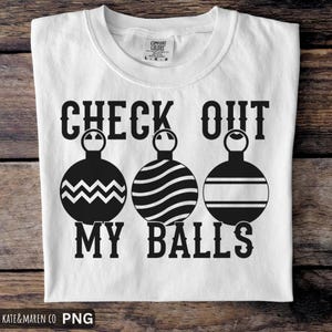 Op de afbeelding: Wit T-shirt met de tekst "CHECK OUT MY BALLS" in dikke zwarte letters. Het ontwerp bevat drie zwarte ornament illustraties met verschillende patronen. Het shirt is gemaakt van een zacht materiaal.