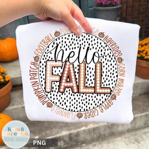 Puede incluir: Una sudadera blanca con un gráfico circular sobre un fondo de puntos negros estilo dálmata. El texto "hello FALL" está centrado en negro y marrón claro. Alrededor del círculo aparecen las palabras "PUMPKIN PATCH", "FOOTBALL", "HAYRIDES", "CORN MAZES", "APPLE CIDER" y "FLANNELS" intercaladas con pequeños iconos de calabazas naranjas.