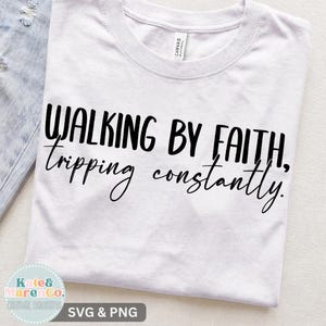 Op de afbeelding: Een wit T-shirt met korte mouwen en de zwarte opdruk Walking by faith, tripping constantly. Het shirt heeft een ronde hals en een standaard pasvorm, waarbij de tekst wordt weergegeven in een combinatie van dikke blokletters en cursief schrift.