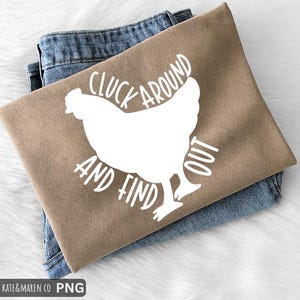 May include: A tan t-shirt with the phrase "CLUCK AROUND AND FIND OUT" in white, along with a white chicken graphic. The shirt is folded and rests on top of a pair of blue jeans.
