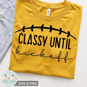 May include: A mustard yellow crew neck t-shirt featuring black text that reads "CLASSY UNTIL kickoff" with a curved line above the text resembling football laces. The shirt has a standard short-sleeve design and a visible white brand label at the neck.