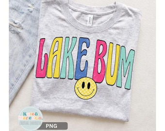 May include: A light gray crew neck t-shirt featuring the text LAKE BUM in large, colorful, distressed-style letters in pink, yellow, blue, and teal. Below the text is a yellow smiley face icon with a checkered pattern and black eyes and mouth.