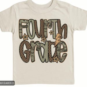 May include: A cream-coloured t-shirt with the words "FOURTH GRADE" in a playful design. The text incorporates camouflage and checkered patterns, with star and lightning bolt accents. The shirt is designed for children.