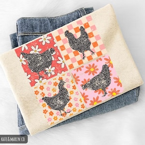 May include: A folded, cream-colored sweatshirt with a colorful chicken design. The design features four squares with a black chicken silhouette on a floral and checkered background. The sweatshirt is resting on a pair of blue jeans.