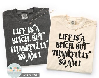 May include: Two t-shirts, one charcoal gray and one cream, with the phrase "LIFE IS A BITCH, BUT THANKFULLY SO AM I" in bold, white lettering. Each shirt has a delicate necklace with a small star pendant.