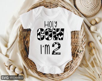 May include: White baby onesie with the text "HOLY COW I'M 2" in black. The words "COW" and the number "2" have a black and white cow print design. The onesie is displayed in a woven basket.