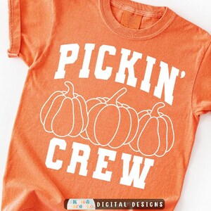 May include: A short-sleeved orange t-shirt featuring white screen-printed text that reads PICKIN' CREW. Below the word PICKIN' are three white line-art illustrations of pumpkins arranged horizontally across the center of the garment.