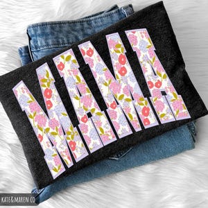K&ouml;nnte beinhalten: Ein dunkelgraues T-Shirt mit dem Wort "MAMA" in gro&szlig;en Buchstaben mit floralem Muster. Die Buchstaben sind mit rosa, roten und lila Blumen gef&uuml;llt. Das T-Shirt liegt gefaltet auf einer Jeans.