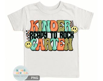 May include: A white short-sleeve t-shirt featuring a graphic design with the text KINDER READY TO ROCK GARTEN. The lettering uses orange, teal, and yellow checkered patterns, accented by two yellow smiley faces and black lightning bolts.