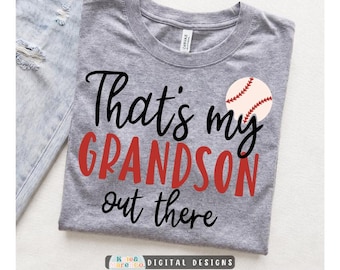 May include: A heather gray t-shirt featuring the black and red text That's my GRANDSON out there with a white baseball graphic containing red stitching. The shirt has a crew neckline and is displayed alongside a piece of light blue denim fabric.