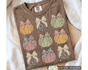 May include: A brown t-shirt featuring a graphic print of nine pumpkins in shades of yellow, green, and pink. Each pumpkin is adorned with a decorative bow in various patterns, including leopard print and checkered designs. The neck label reads Comfort Colors.
