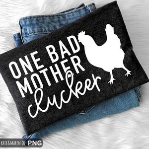 Op de afbeelding: Een donkergrijs t-shirt met de witte tekst "ONE BAD MOTHER clucker" en een witte kippengrafiek. Het t-shirt is opgevouwen op een blauwe jeans, op een witte, zachte ondergrond.
