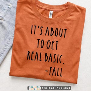 May include: A burnt orange short-sleeved t-shirt with black text printed on the front that reads: IT'S ABOUT TO GET REAL BASIC. -FALL. The shirt has a crew neckline and is displayed alongside a piece of light blue distressed denim fabric.