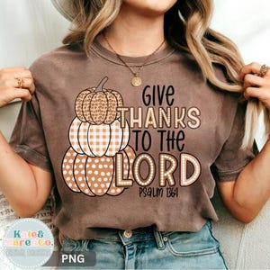 May include: A brown short-sleeve t-shirt featuring a graphic of three stacked pumpkins with leopard, plaid, and polka dot patterns. The text reads "GIVE THANKS TO THE LORD PSALM 136:1" in black and light brown lettering with a dotted outline effect.