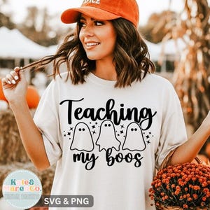 May include: A cream-colored short-sleeve t-shirt featuring black text that reads Teaching my boos, accompanied by a graphic of three simple ghost outlines and small star shapes printed on the front center of the garment.