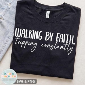 Op de afbeelding: Een zwart t-shirt met korte mouwen en de witte opdruk Walking by faith, tripping constantly. Het shirt heeft een ronde hals en ligt naast een lichtblauwe, versleten spijkerstof met zichtbare gerafelde randen aan de zijkant.