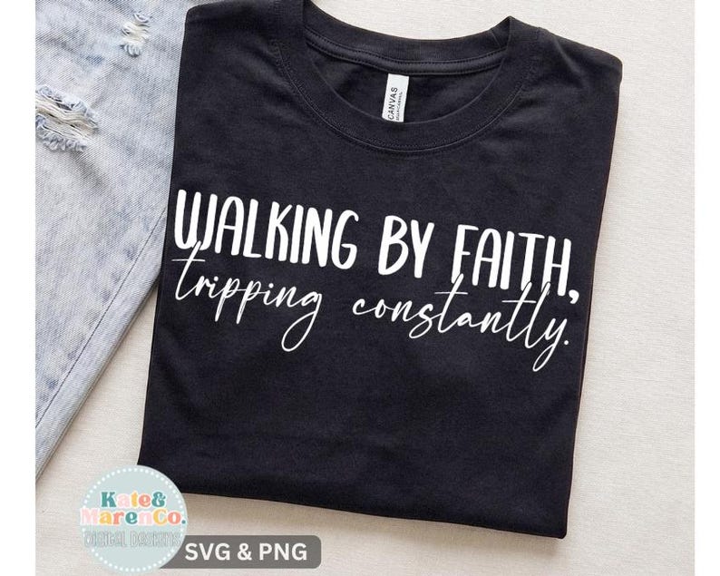 Op de afbeelding: Een zwart t-shirt met korte mouwen en de witte opdruk Walking by faith, tripping constantly. Het shirt heeft een ronde hals en ligt naast een lichtblauwe, versleten spijkerstof met zichtbare gerafelde randen aan de zijkant.