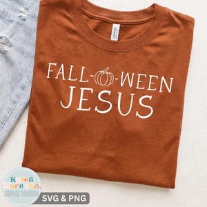 May include: A burnt orange short-sleeved t-shirt featuring white text that reads FALL-WEEN JESUS, with a simple line-art pumpkin graphic replacing the hyphen. The shirt is displayed alongside a piece of light blue distressed denim fabric.