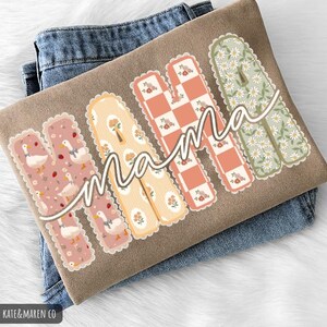 May include: A tan t-shirt featuring the word MAMA in large, scalloped-edge letters. Each letter has a unique pattern: ducks on pink, yellow stripes with flowers, a peach and white checkerboard, and white daisies on a sage green background.