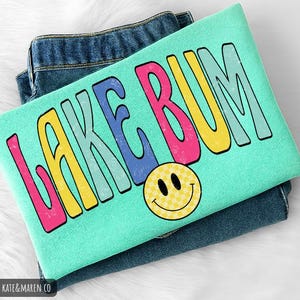 May include: A mint green t-shirt featuring the text LAKE BUM in large, colorful, distressed-style letters in pink, yellow, blue, and light green. Below the text is a yellow smiley face with a checkered pattern. The shirt is folded over blue jeans.