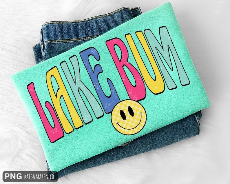 May include: A mint green t-shirt featuring the text LAKE BUM in large, colorful, distressed-style letters in pink, yellow, blue, and light green. Below the text is a yellow smiley face with a checkered pattern. The shirt is folded over blue jeans.