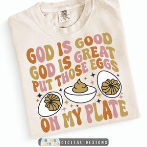 May include: A cream-colored t-shirt featuring the text GOD IS GOOD GOD IS GREAT PUT THOSE EGGS ON MY PLATE in retro-style orange, pink, and brown lettering, accompanied by illustrations of three deviled eggs and small black star accents.