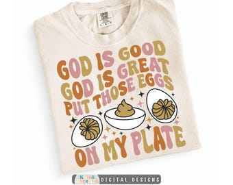 May include: A cream-coloured t-shirt featuring the text GOD IS GOOD GOD IS GREAT PUT THOSE EGGS ON MY PLATE in retro-style orange, pink, and brown lettering, accompanied by illustrations of three deviled eggs and small black star accents.