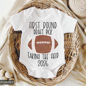Könnte beinhalten: Weißer Baby-Body mit dem Text "FIRST ROUND DRAFT PICK TAKING THE FIELD 2026" und einer braunen Football-Grafik. Präsentiert in einem geflochtenen Korb mit dekorativen Elementen.