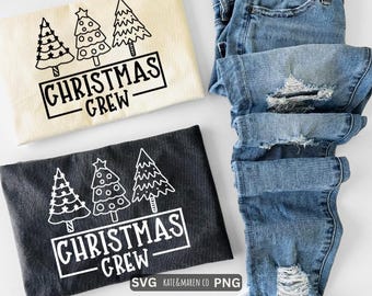 May include: Two folded t-shirts with a Christmas theme, featuring three Christmas tree designs and the text "CHRISTMAS CREW" in a rectangular frame. The shirts are cream and dark gray. A pair of blue jeans is folded next to the shirts.