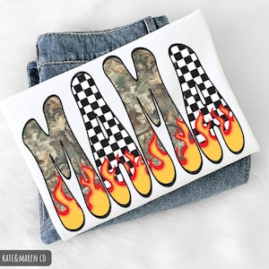 May include: White graphic tee with the word "MAMA" in a unique design. The letters feature a camouflage pattern, checkered flag, and flames. The tee is laid on top of a pair of blue jeans.