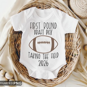 Puede incluir: Body de bebé blanco con el texto "FIRST ROUND DRAFT PICK" sobre un gráfico de fútbol americano marrón y "TAKING THE FIELD 2026" debajo. Presentado en una cesta de mimbre con elementos decorativos.
