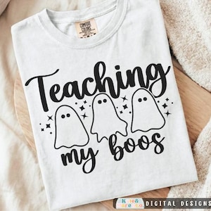 May include: A white short-sleeved t-shirt featuring a black graphic print of three ghosts and the text "Teaching my boos" with small star accents. The shirt has a crew neckline and a Comfort Colors brand label visible on the inside collar.