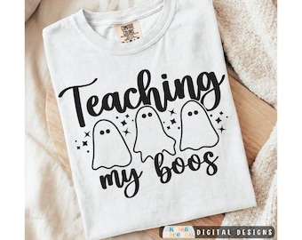 May include: A white short-sleeved t-shirt featuring a black graphic print of three ghosts and the text "Teaching my boos" with small star accents. The shirt has a crew neckline and a Comfort Colors brand label visible on the inside collar.