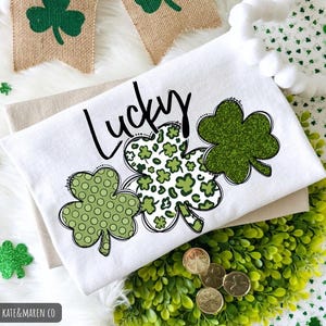 K&ouml;nnte beinhalten: Wei&szlig;es Handtuch zum Thema St. Patrick's Day mit dem Wort "Lucky" und drei Kleeblatt-Designs. Ein Kleeblatt hat ein glitzerndes gr&uuml;nes Finish, ein anderes hat ein Tupfenmuster und das dritte hat ein Leopardenmuster. Das Handtuch ist auf einem beigen Tuch gefaltet.