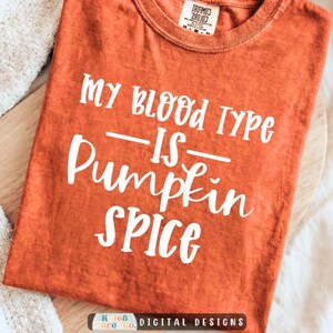May include: A burnt orange short-sleeve t-shirt with a crew neckline. The shirt features white text printed on the front that reads, "MY BLOOD TYPE IS PUMPKIN SPICE." The fabric has a textured, washed appearance with a Comfort Colors brand label.