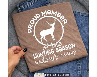 Pode incluir: Uma t-shirt castanha com um gráfico branco de um veado dentro de um círculo e o texto PROUD MEMBER OF THE HUNTING SEASON widow's club em letras brancas. A t-shirt está dobrada e colocada sobre um par de calças de ganga azuis.