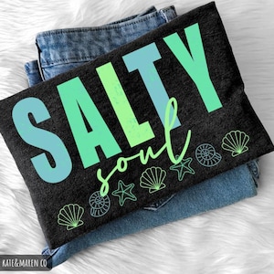 Pu&ograve; includere: Una maglietta nera con la scritta "SALTY soul" in blu e verde, con grafiche di conchiglie e stelle marine. La maglietta &egrave; piegata sopra un paio di jeans blu, appoggiata su una superficie bianca e soffice.