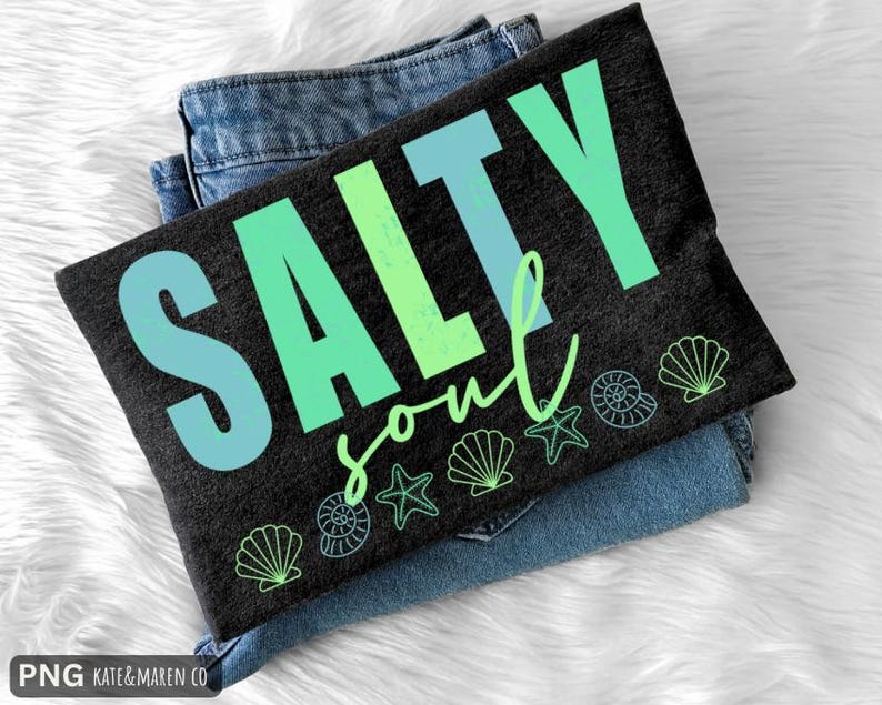 Pu&ograve; includere: Una maglietta nera con la scritta "SALTY soul" in blu e verde, con grafiche di conchiglie e stelle marine. La maglietta &egrave; piegata sopra un paio di jeans blu, appoggiata su una superficie bianca e soffice.