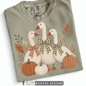 May include: A sage green t-shirt featuring a graphic of three white geese wearing plaid bows, surrounded by orange and white pumpkins and autumn leaves. The shirt has a Comfort Colors brand label at the neck, indicating a size large.